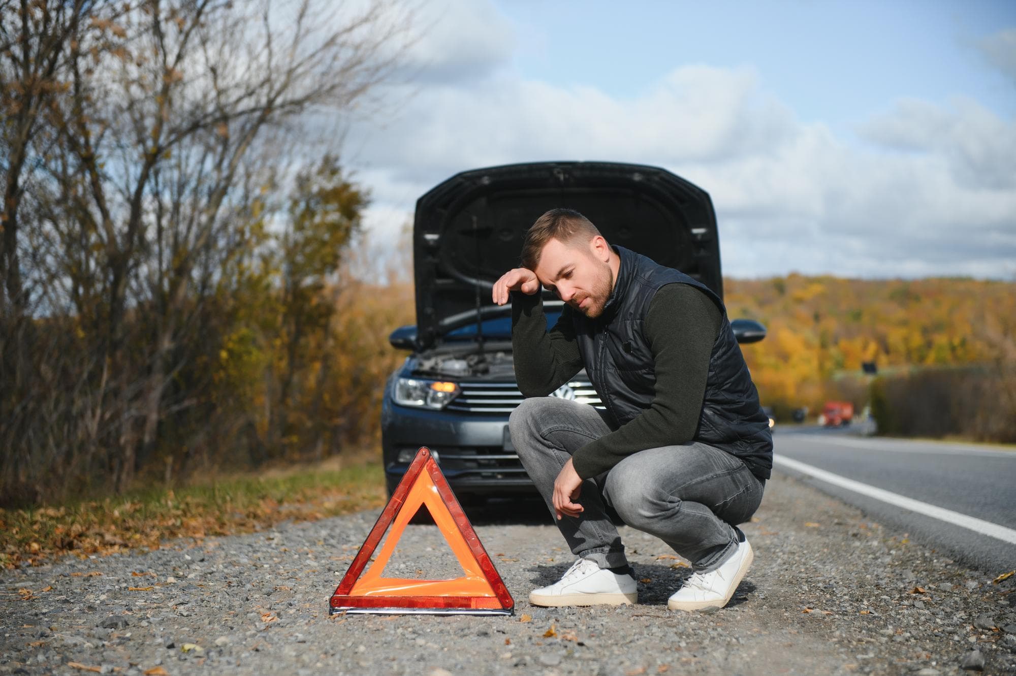 Frustrated driver next to car with hood open on roadside, warning triangle placed in front for safety